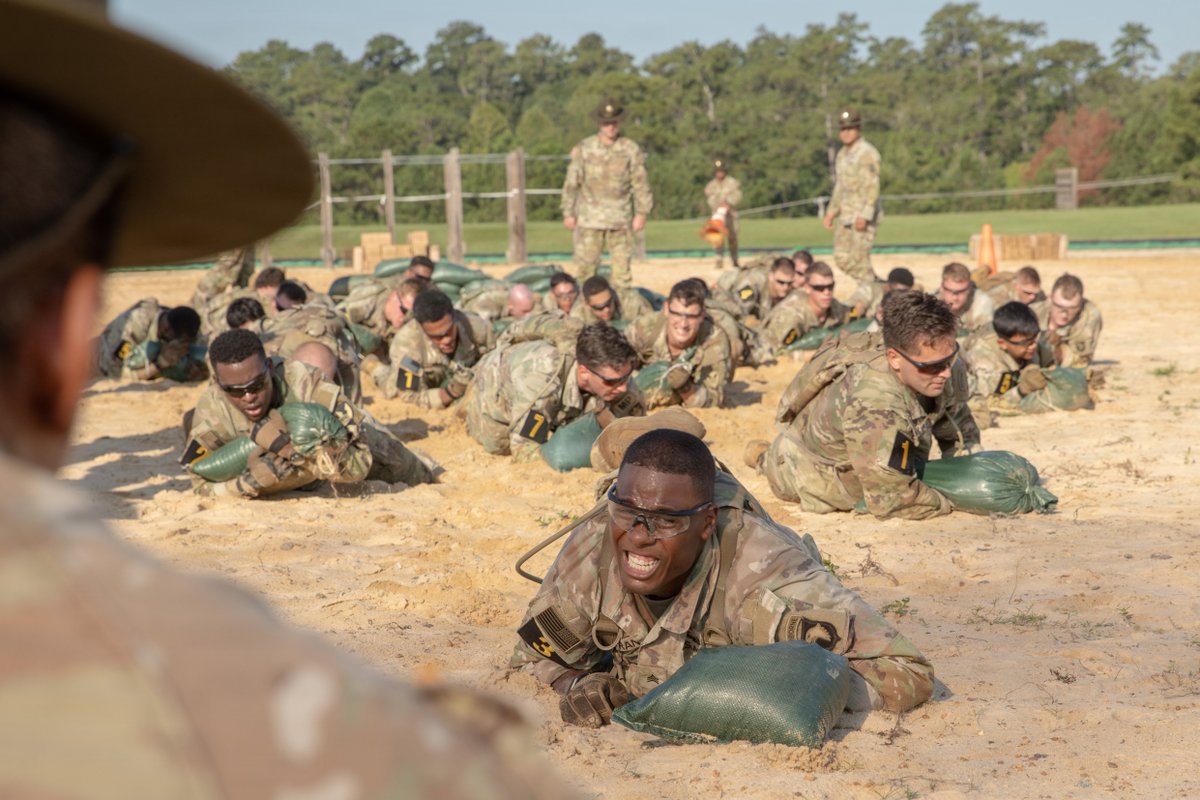 Soldiers in camouflage uniforms crawl through deep sand, pushing green sandbags. An African American soldier in the foreground grimaces with intense effort, wearing safety glasses. Other soldiers behind him also push bags. Instructors in drill sergeant hats observe from the background.
