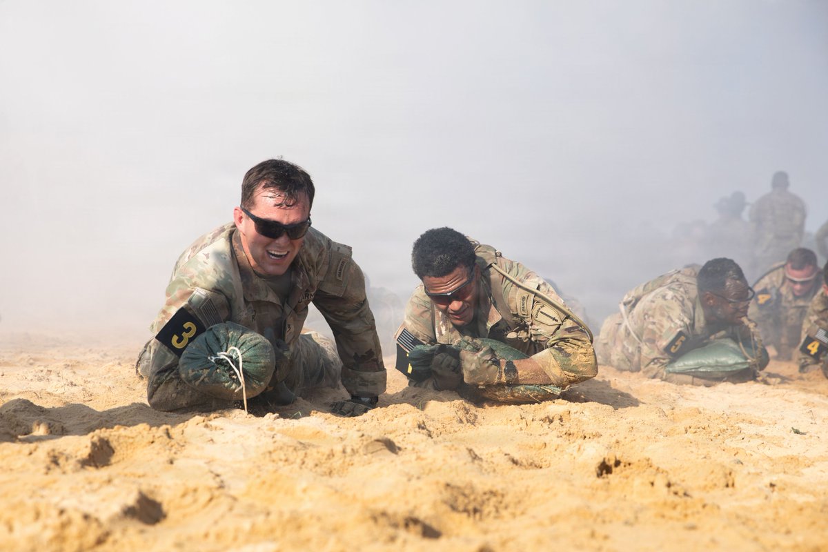 Two soldiers in camouflage uniforms and sunglasses low-crawl across sandy terrain, pushing green sandbags. The left soldier, with a '3' patch, grimaces in effort. The right soldier, wearing 'RANGER' and 'FOLLOW ME' patches, also strains. Other blurred soldiers crawl in the smoky background.