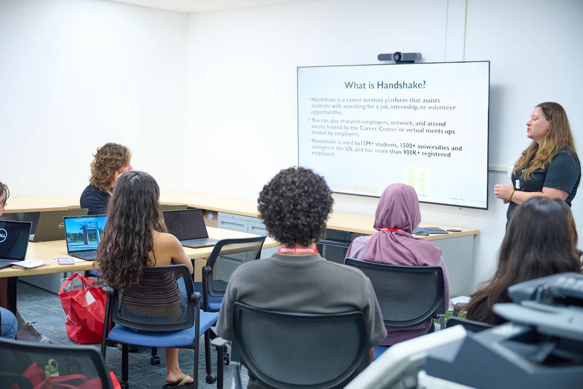 A woman presents a slide about Handshake to a classroom of attentive students seated with laptops.