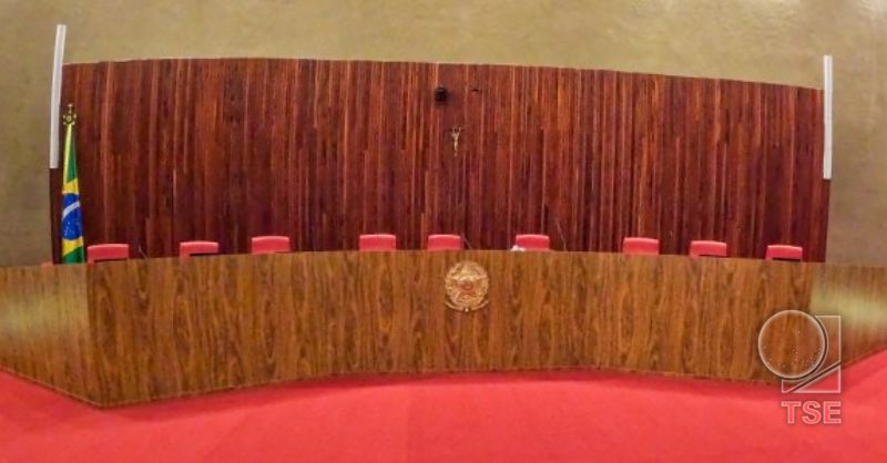 Sala plenária do TSE vazia, com longa mesa de madeira curva à frente, brasão da República no centro e cadeiras vermelhas atrás. Ao fundo, parede de madeira com crucifixo e bandeira do Brasil à esquerda. Piso vermelho e marca d’água do TSE.