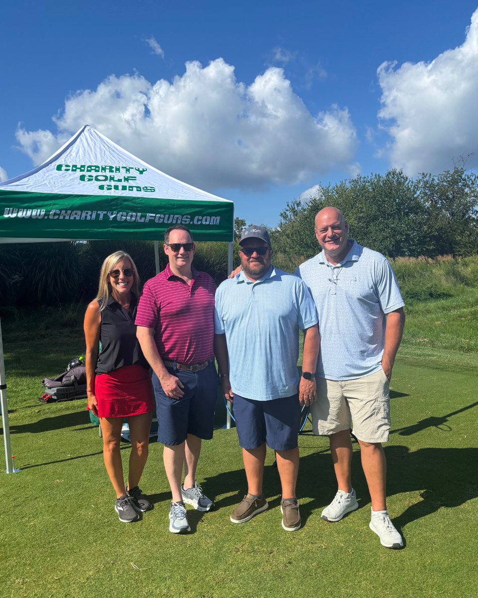 Four people standing on a grassy area in front of a white tent with "Charity Golf Guns" written on it. They are dressed in casual golf attire, and the sky is partly cloudy.
