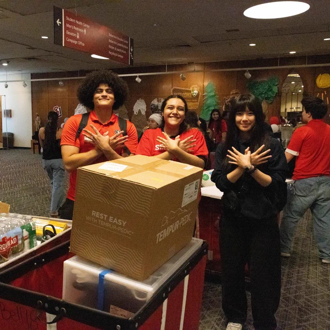 Three Seattle University students stand behind a red moving cart in Bellarmine Hall full of items for a dorm room.