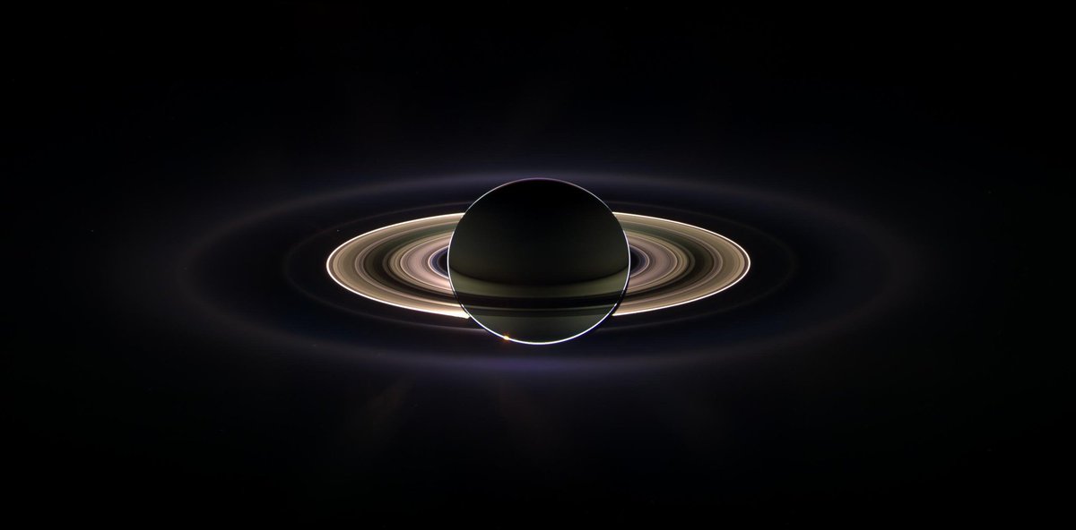 A striking image of Saturn, shown in silhouette, with its rings illuminated against the dark backdrop of space. The sunlight creates a halo effect around the planet and its rings.