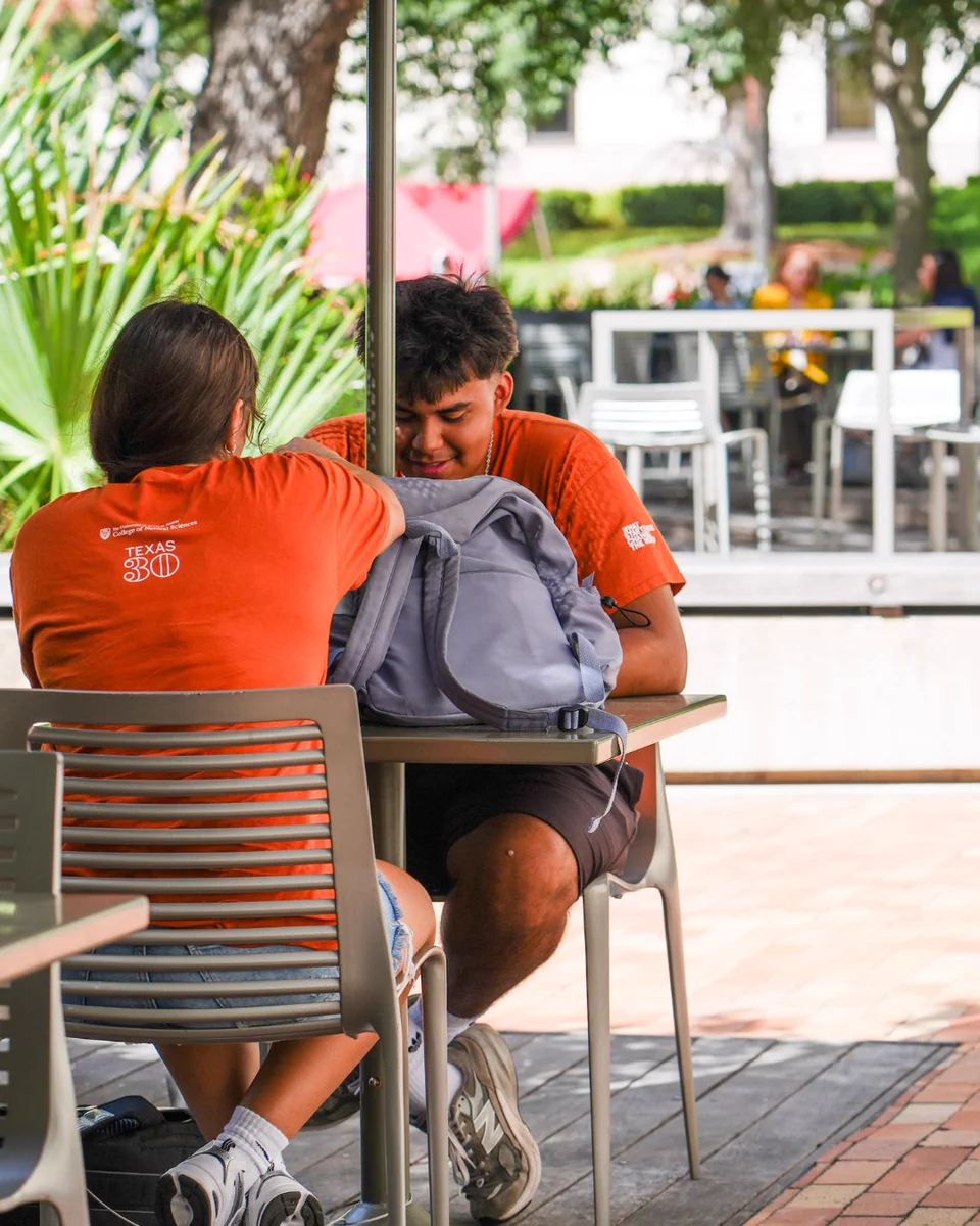 Two young people in orange shirts sit at an outdoor table. The person on the left wears a shirt with \"TEXAS 30\" on the back. The other person looks down at a gray backpack on the table between them. Green foliage is visible in the background.