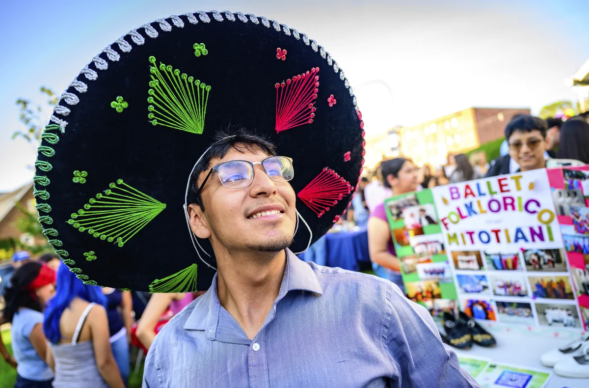 A student wearing a large, colorful sombrero and glasses stands smiling outdoors. In the background, there is a display board titled "Ballet Folklorico Mitotiani," along with several people interacting at an event. The atmosphere is bright and festive.