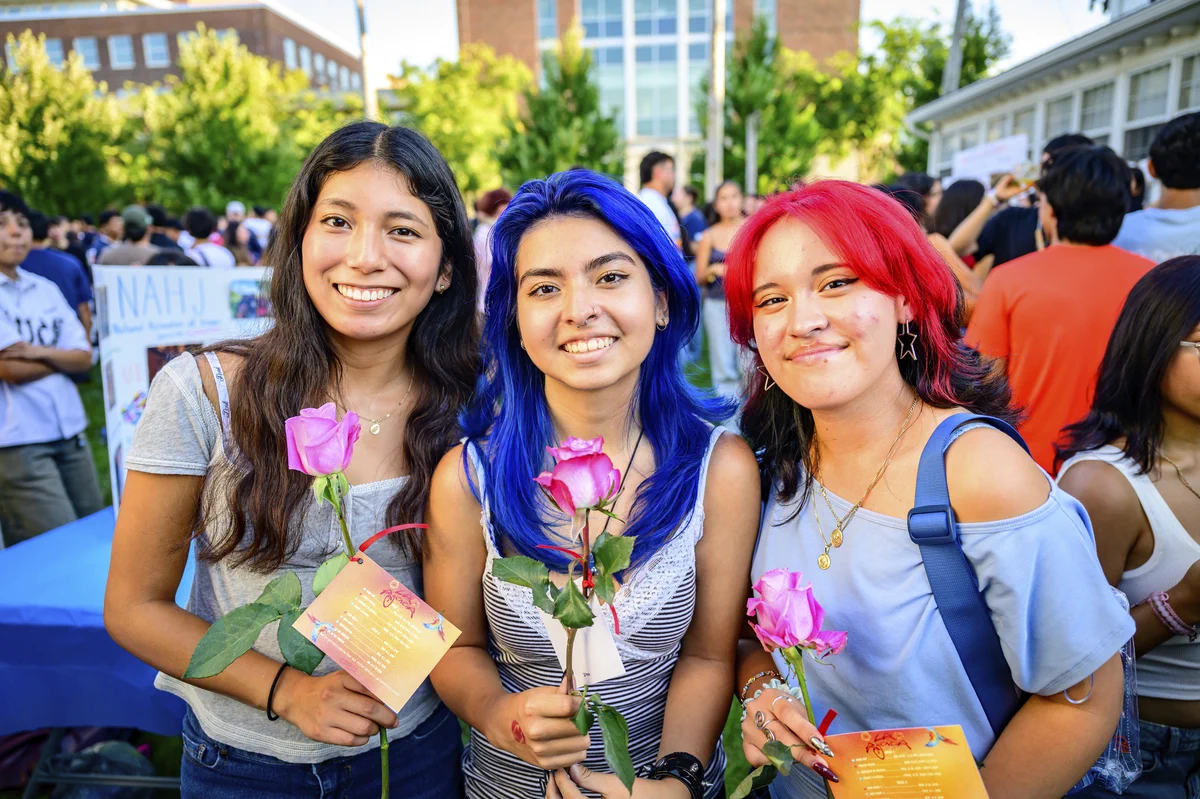 Three people are smiling and holding pink roses with cards at an outdoor event. A building and other attendees are visible in the background.