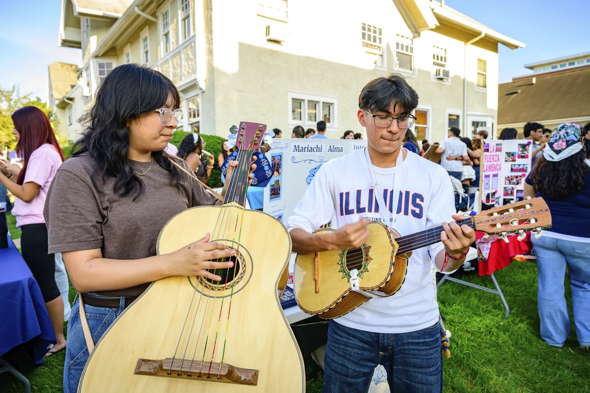 Two individuals are playing traditional stringed instruments outdoors at an event. One is strumming a large vihuela, and the other is playing a smaller guitar-like instrument. The background shows informational displays and a group of people mingling nearby.