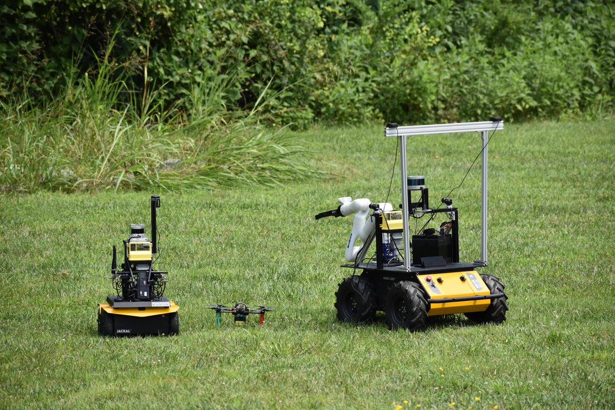 Three robotic explorers in a grassy field: a drone parked between two rovers. The larger rover has a multi-jointed arm extending from it.