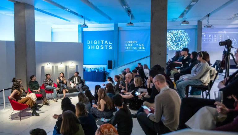 Photo of a panel discussion at a "Digital Ghosts" event. Four panelists sit in red and gray chairs against a wall with projected text reading "Digital Ghosts" and "Dorsey, Kaufmann & Feeling Data." A large seated audience fills the foreground, with blue lighting throughout the gallery-like space.