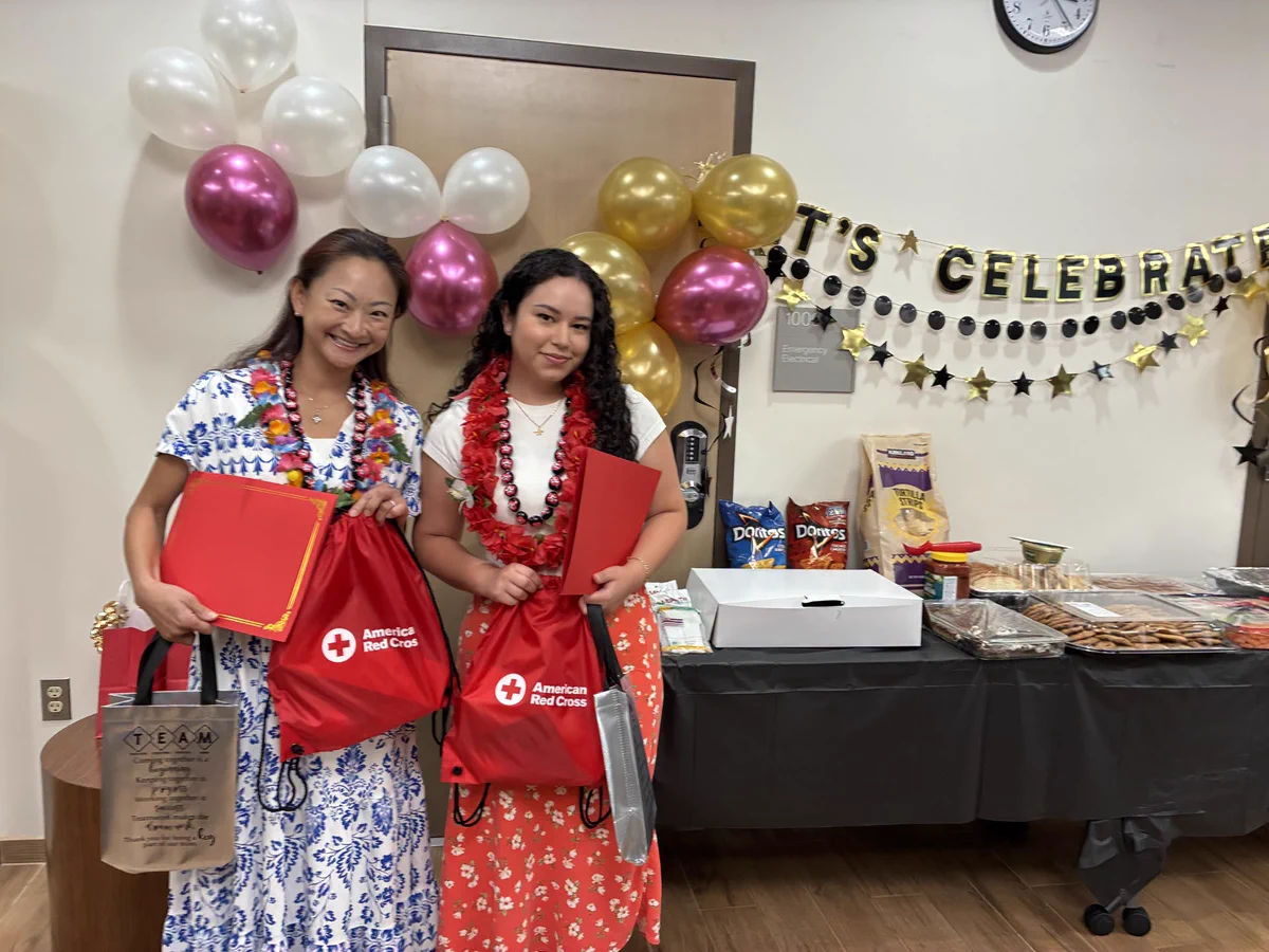 Two people stand holding American Red Cross bags, surrounded by festive decorations, including balloons and a “Let's Celebrate” banner. A table nearby is filled with snacks and drinks.