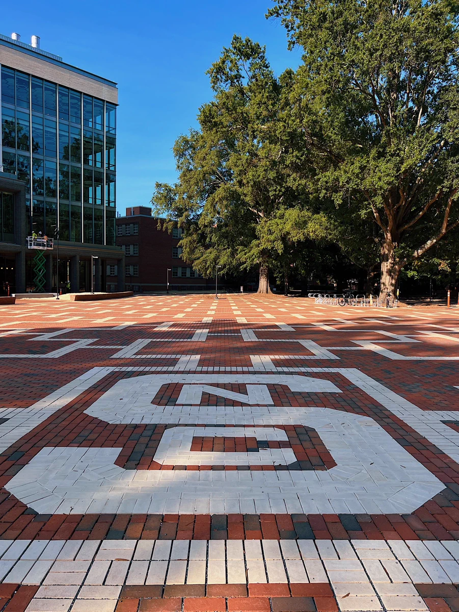 Large brick courtyard with a white painted 'NCSU' logo in the center, surrounded by tall green trees and a modern glass building on the left.
