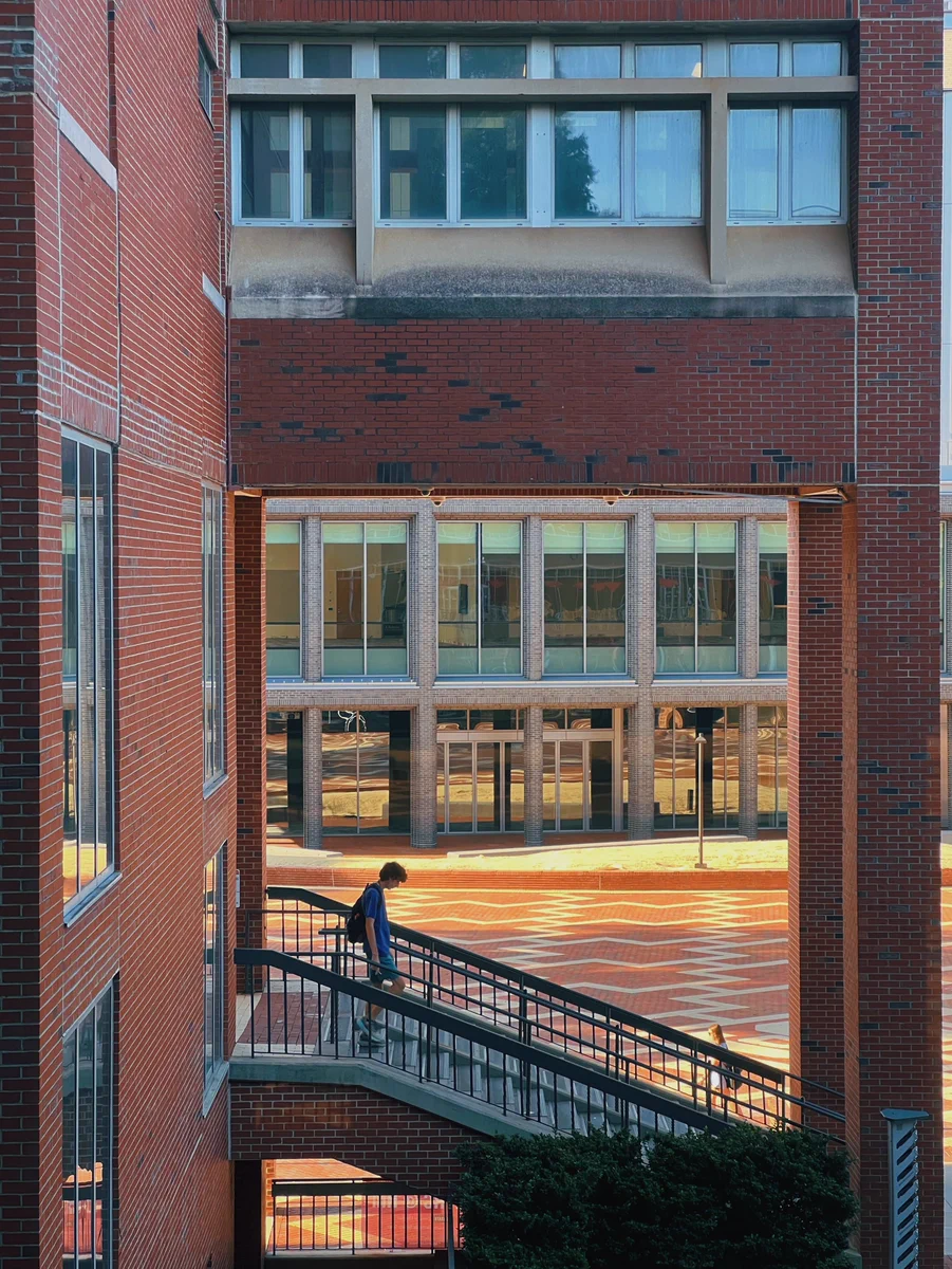 Person walking down a ramp between two red brick buildings with large windows reflecting sunlight.