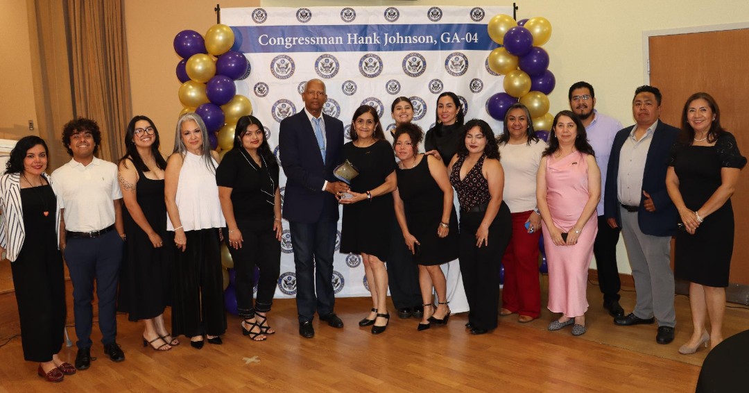 A group of adults pose for a photo in front of a decorated event backdrop, with purple and gold balloons. A man and woman near the center hold a small award. There is text on the backdrop.
