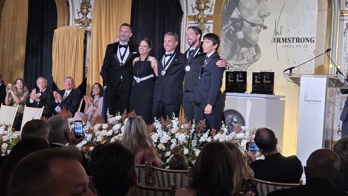A group of elegantly dressed people posing on a stage at the Armstrong Space Prize event, flanked by a seated audience. Several items are displayed on a table.