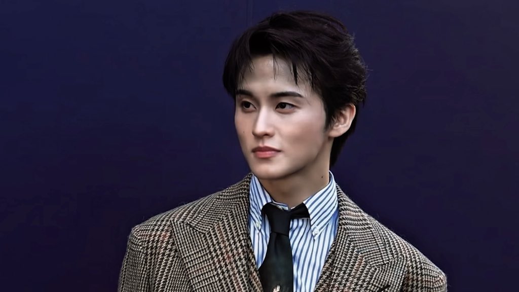 A young man with dark, tousled hair poses against a deep purple backdrop, wearing a tailored houndstooth blazer over a blue and white striped button-down shirt paired with a black necktie—his polished, high-fashion ensemble reflecting the "expensive as hell" caption.