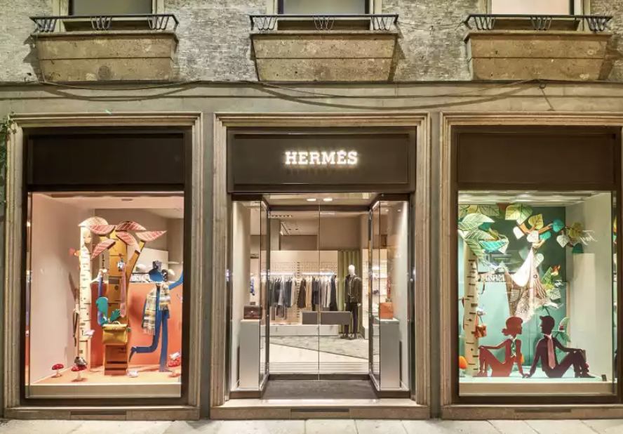 An Hermès boutique storefront photographed at night, with the illuminated Hermès wordmark in white serif letters above a glass double door set into a stone facade with iron balconies overhead. Two large display windows flank the entrance, each dressed with whimsical paper-cut installations: on the left, a stylised birch tree with a climbing figure in a striped scarf and a mannequin in a tan coat beside red-and-white toadstools; on the right, a green woodland scene with cut-out leaves, a hanging chair, and two silhouetted figures seated back to back on the forest floor. Through the open door, warm-lit interior shelves of leather goods and ready-to-wear are visible with a lone mannequin. The image accompanies an Intern Pierre research note on Hermès allocation, the Birkin game, legacy client churn and the secondary market.