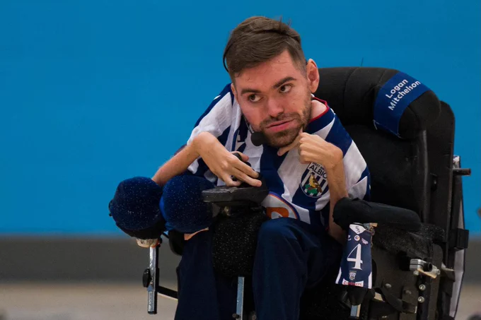 Harrison, wearing a blue-and-white striped West Bromwich Albion Powerchair Football kit, controls a powerchair during an indoor match, with a player number card attached to the chair.