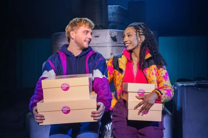 Sam Tutty and Christiani Pitts in Two Strangers, they hold boxes and smile at each other