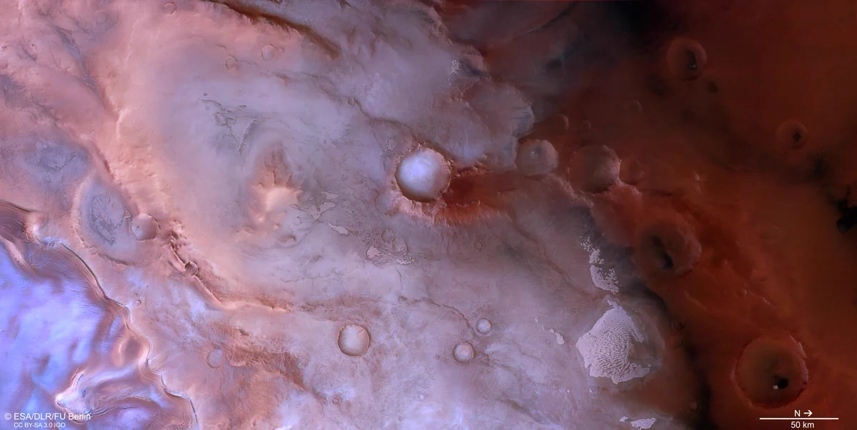 A purple-pink-coloured patch of Mars, which is lighter on the left and darker on the right. A dark line runs from the top centre towards the bottom right, dividing the light and dark areas. At the centre of the image is a sharp light-coloured crater. Less sharp craters are scattered over the rest of the image. At the bottom left is a bright white-blue triangular patch, it looks icy. A small scale bar at the bottom right indicates that north is to the right, and that a line approximately one tenth of the image width is 50 km long.