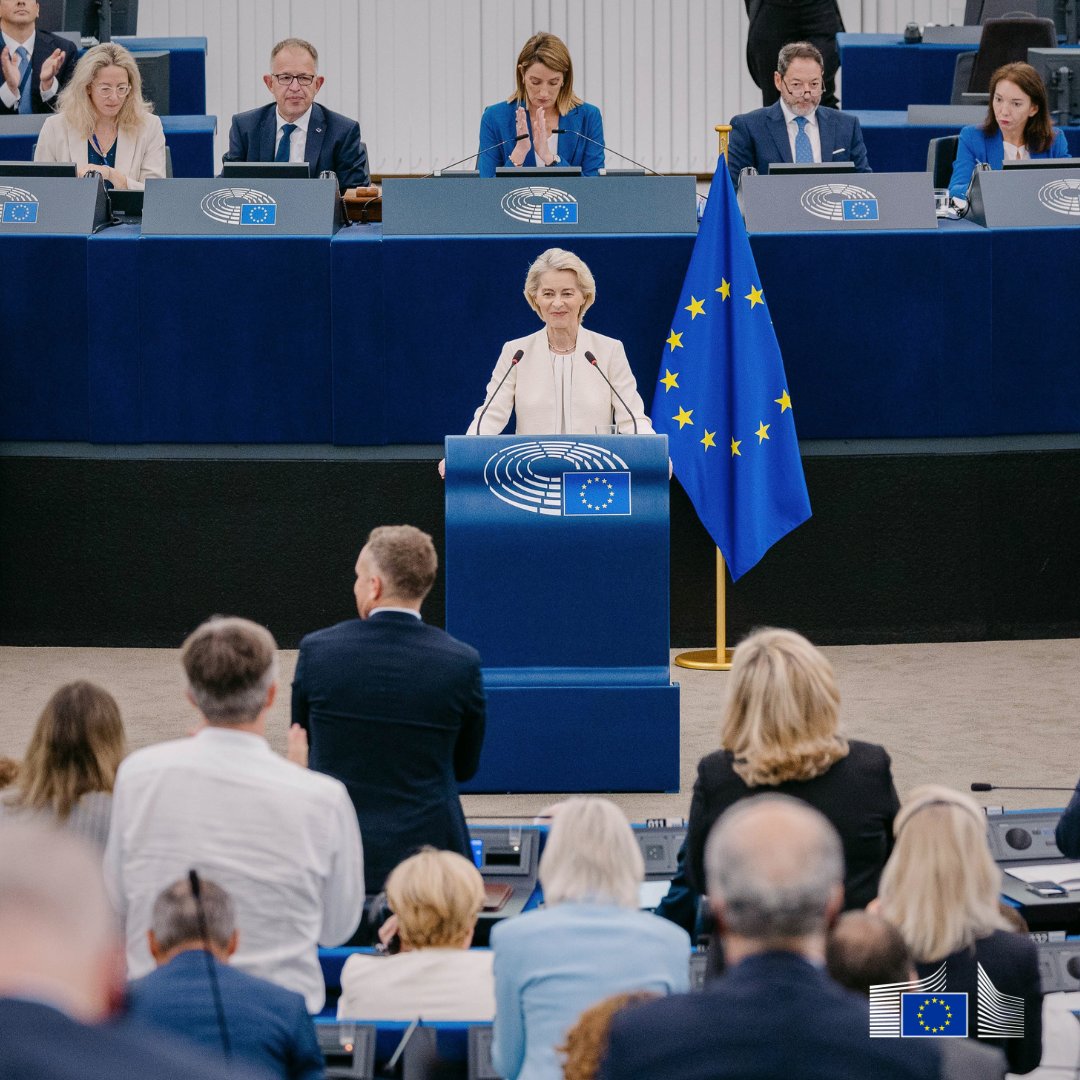 European Commission President Ursula von der Leyen, wearing a light-colored suit, speaks from a blue podium. To her right stands the European Union flag. Seated on a raised platform behind her is European Parliament President Roberta Metsola along with other officials, while members of parliament watch from the foreground.