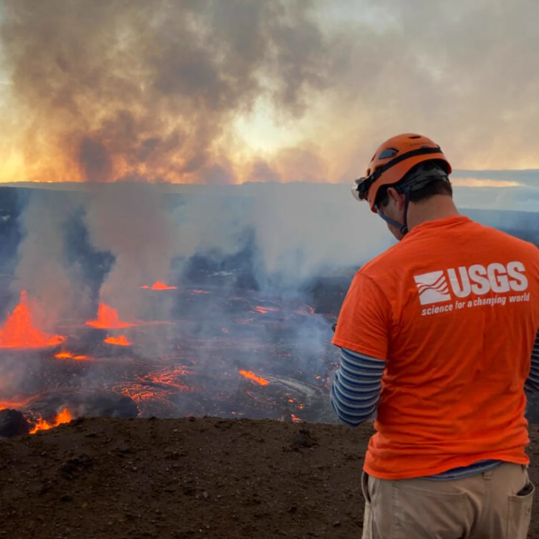 USGS scientist wearing an orange helmet and shirt observing an active volcanic lava flow emitting smoke and glowing molten rock.