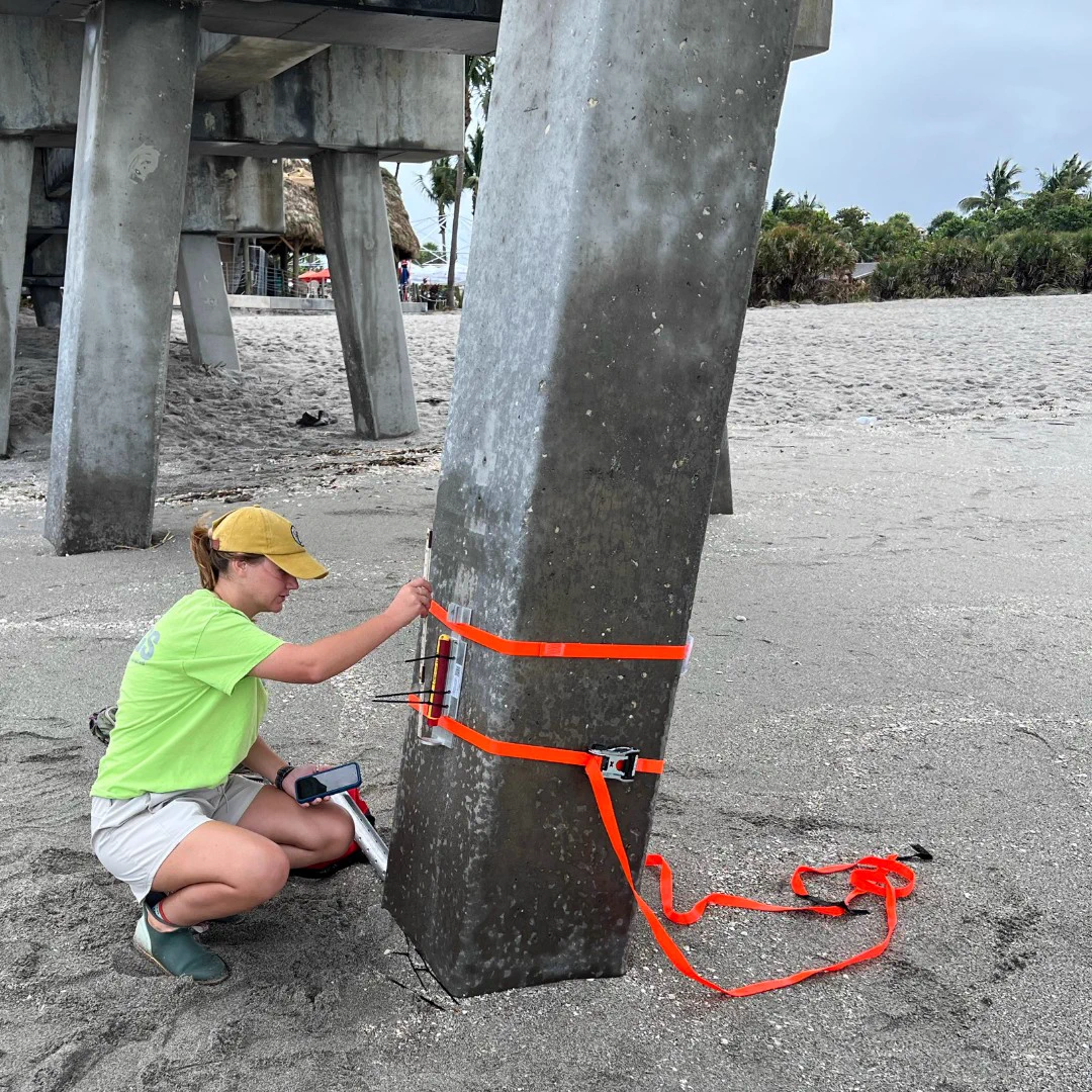 Person in a yellow cap and green shirt securing orange straps around a concrete pier leg on a sandy beach.