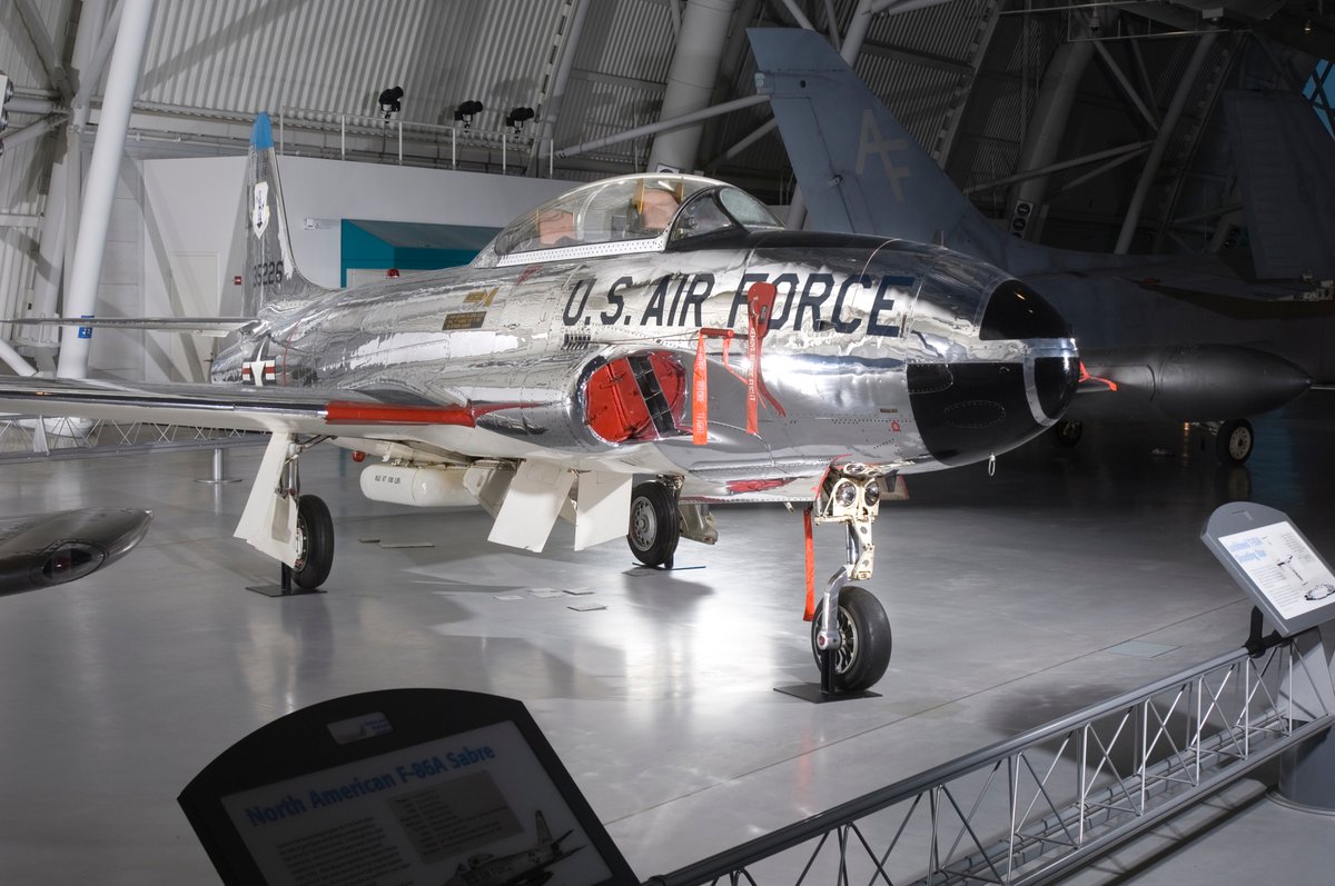 A jet displayed in a museum setting. The aircraft is polished and silver, featuring "U.S. Air Force" on its side. Various informational signs and displays are visible around the jet.