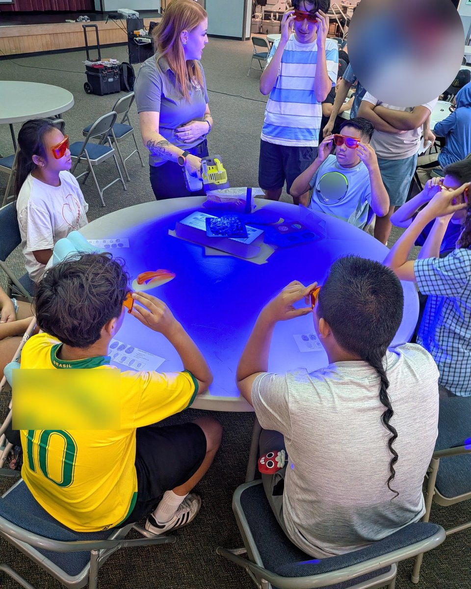 Children wearing 3D glasses gathered around a round table with a glowing blue surface, guided by an adult instructor in a classroom setting.