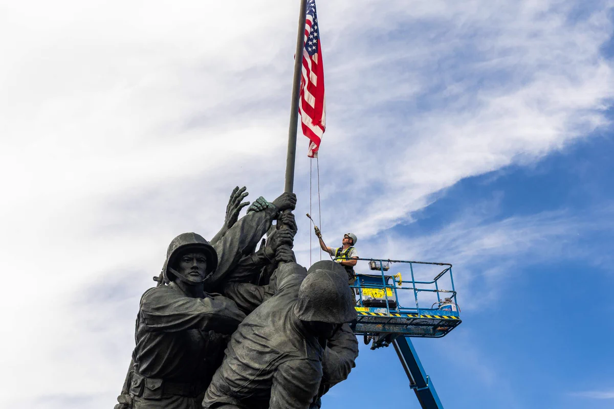 Workers cleaning the Iwo Jima Memorial statue with an American flag against a partly cloudy sky.