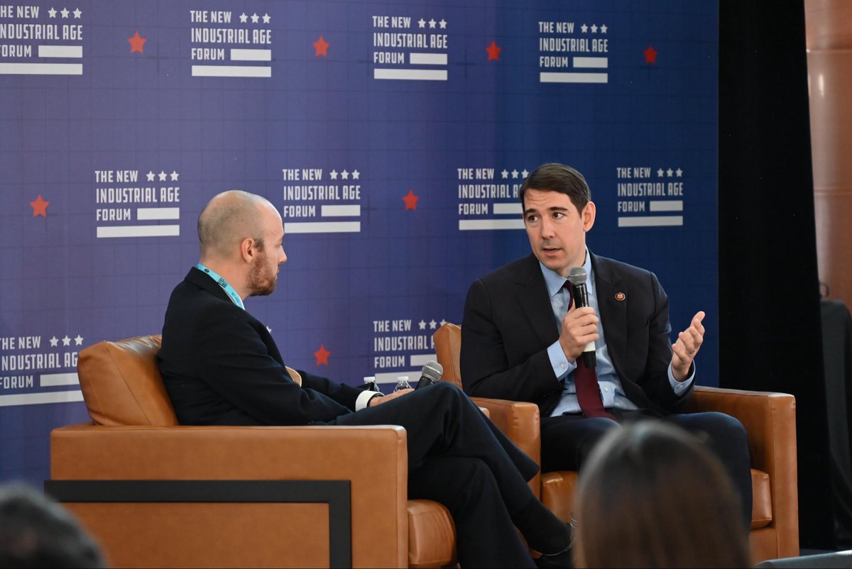 Two men in suits sit in armchairs during a live discussion. One man speaks into a handheld microphone while gesturing; the other faces him and listens. A branded event backdrop is behind them, with audience members partly visible in the foreground.