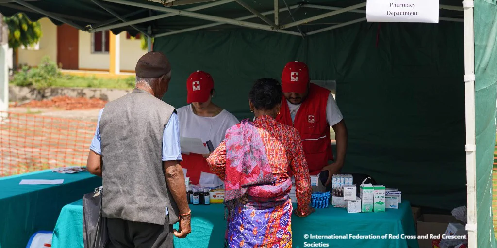 Two Red Cross workers assist individuals at an outdoor medical aid booth with medicines on display.