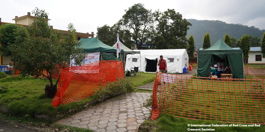 Temporary medical tents set up outdoors with a person in a red vest walking near orange safety fencing on a cloudy day.