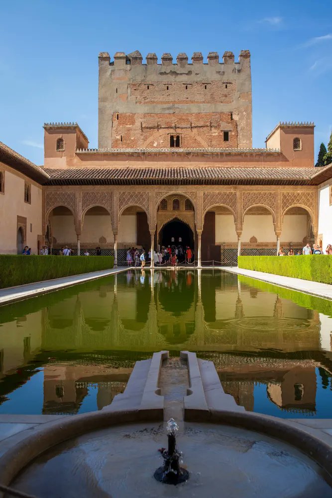 Photo shows the Court of the Myrtles (Patio de los Arrayanes), the central courtyard of the Comares Palace located within the historic Alhambra complex in Granada, Spain. A medieval Moorish brick palace looms over the long water feature and fountain.
Image courtesy of thenotsoinnocentsabroad.com