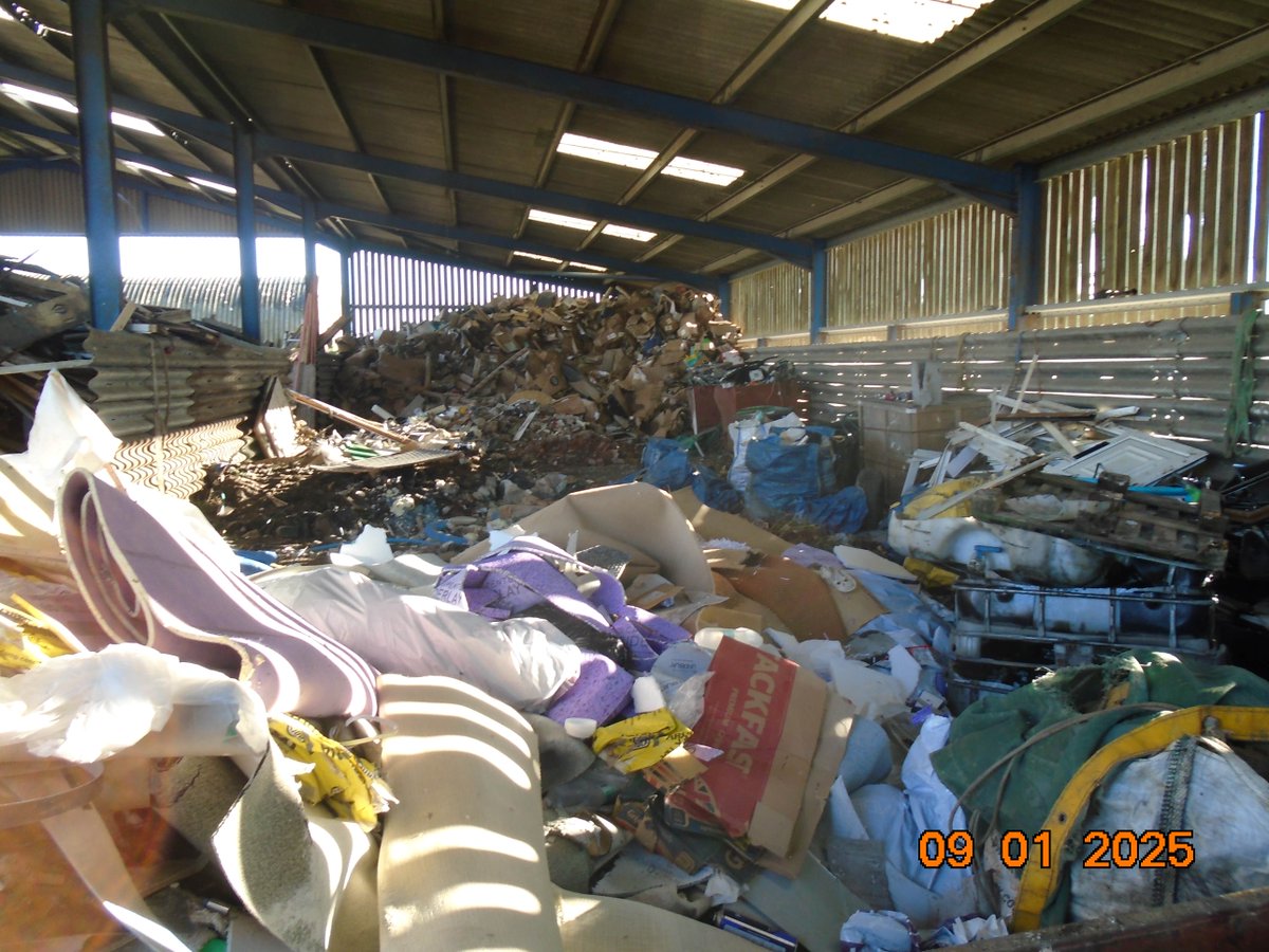 A large indoor area filled with various piles of debris and discarded materials, including fabrics, cardboard, and metal objects. Sunlight filters through the partially open sides of the structure. A stack of wooden materials is visible in the background.