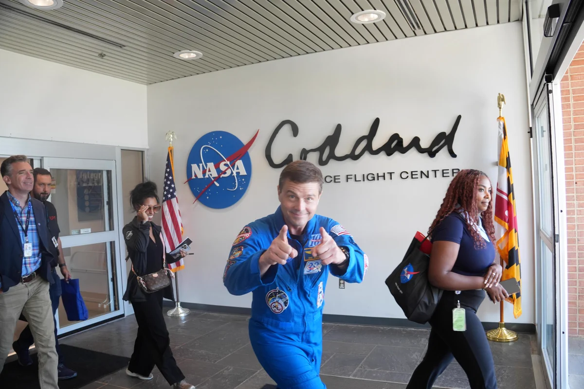 Reid Wiseman exiting a building through sliding glass doors. Reid has a beaming smile on his face, hunching down to point enthusiastically at the camera. Behind him is a big Goddard logo, a U.S. flag, and a Maryland flag. 