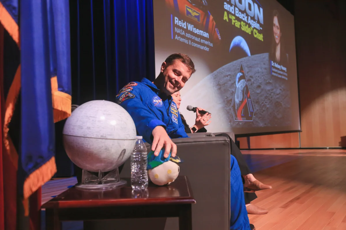 Reid Wiseman sitting on a stage, taken from the side. He is looking down in the direction of the camera, at a small Rise zero gravity indicator, which he is picking up. A Moon globe sits next to Rise on the table. 