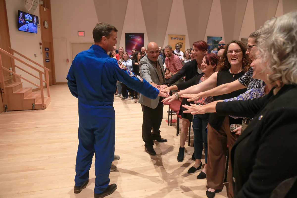 Reid Wiseman putting his hand in with a crowd of Goddard employees, all reaching toward the center. They are all standing up and smiling. Many people are visible reaching toward the hands in spot in the background. 