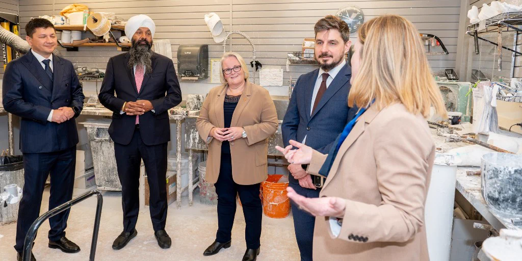 Five people in business attire engaged in a discussion inside a workshop filled with tools and materials.
