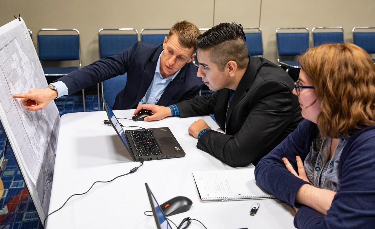 A person points at a large printed document on a table, showing it to two others. The group is gathered around a laptop and a notebook in a conference room with blue chairs and a beige wall.