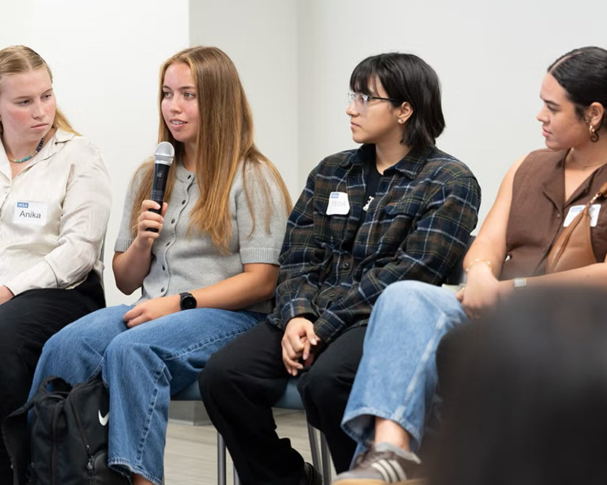 Four students sit together during a group discussion as one student speaks into a microphone and the others look toward her.