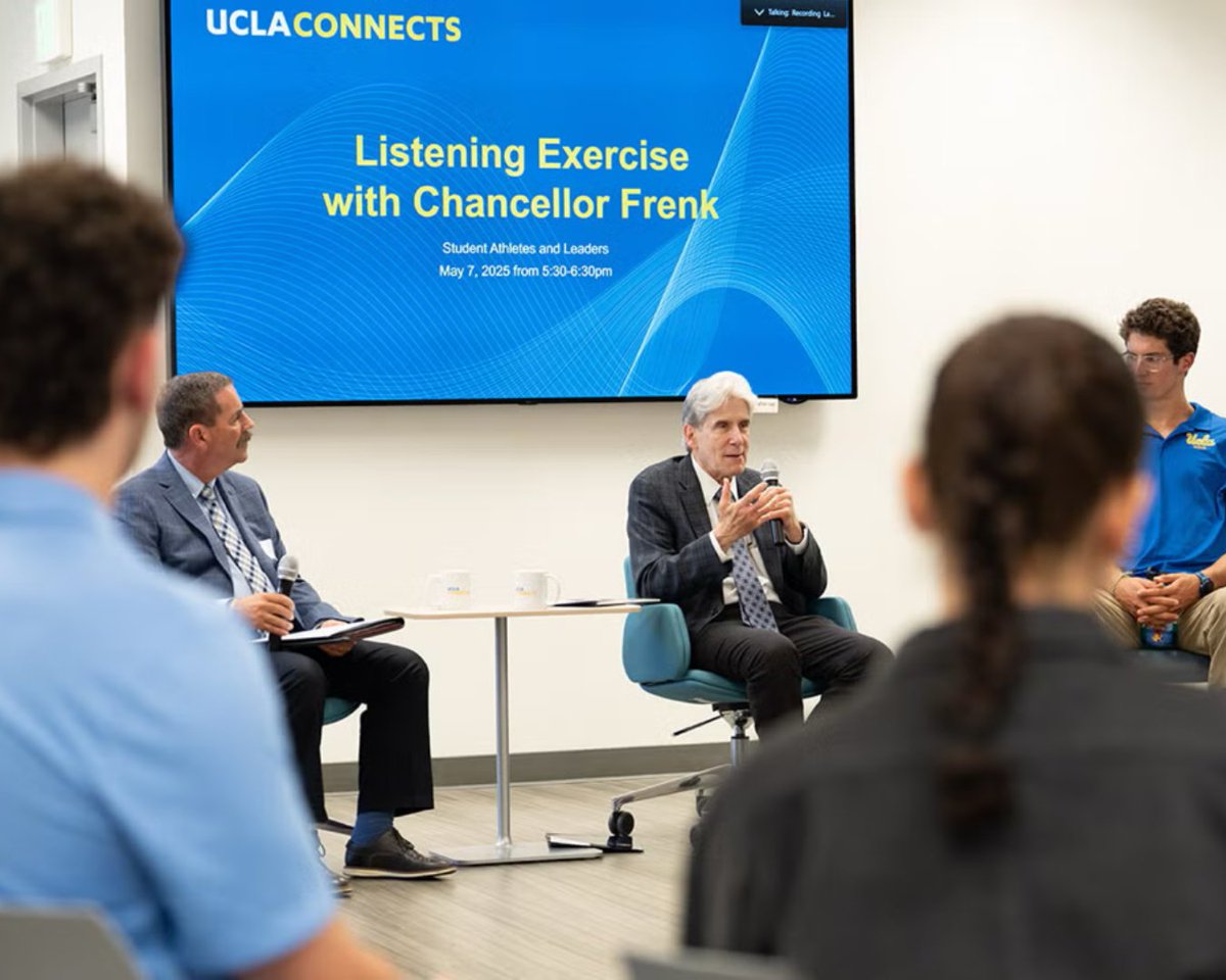 A gray-haired man in a suit sits at the front of a room speaking into a microphone, with students seated around him and a screen behind him reading “UCLA Connects: Listening Exercise with Chancellor Frenk.”