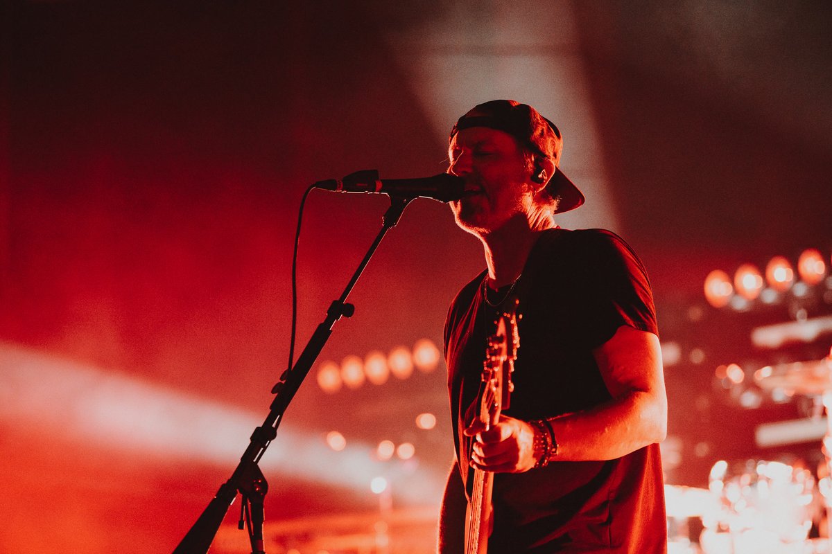 Musician playing guitar and singing into a microphone under red stage lights.