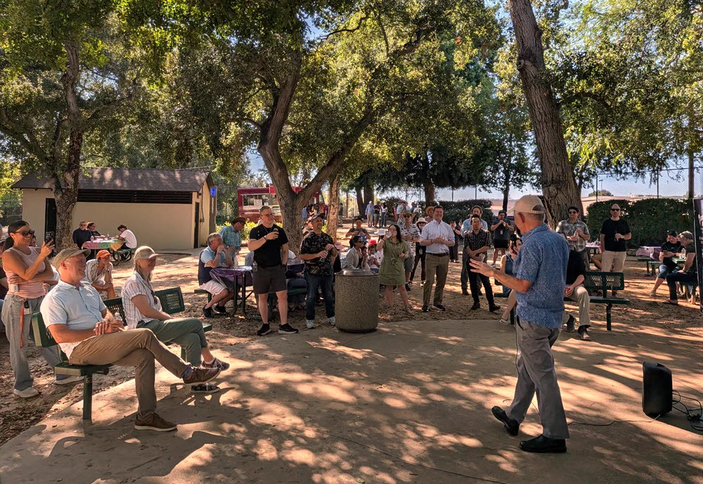 A group of about 30 people are gathered in a park under a shady canopy of oaks, either standing or sitting on benches, while listening to IPAC Executive Director Tom Greene, who is standing and facing the crowd.