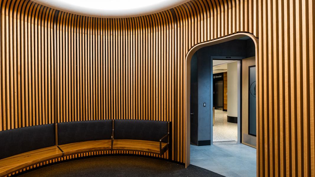 Curved wood-slat lobby with a semicircular upholstered bench and an open doorway leading to an elevator area.