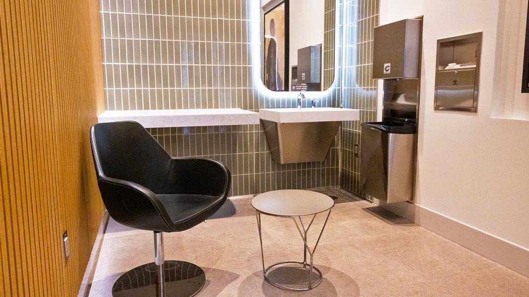 Modern tiled washroom with a backlit mirror, wall-mounted sink, black lounge chair, small round table, and metal fixtures.