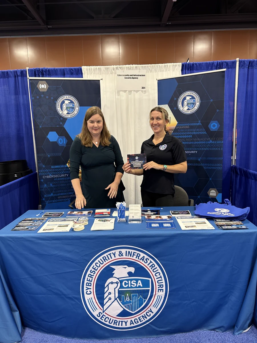 Booth display featuring CISA (Cybersecurity and Infrastructure Security Agency) logos and informational materials. Two individuals stand behind the table, one holding a book. The setup includes banners and promotional items related to cybersecurity.