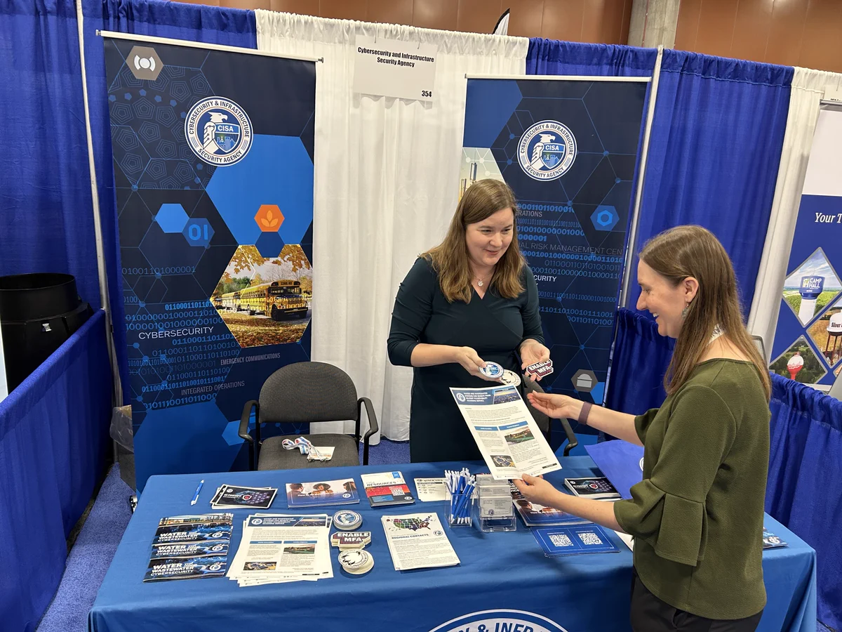 A person is standing at a table displaying informational materials at a convention booth. Another individual, across the table, is handing a pamphlet to the person at the booth. The table is covered with brochures, pens, and a small bowl of items. The backdrop features CISA logos and graphics.