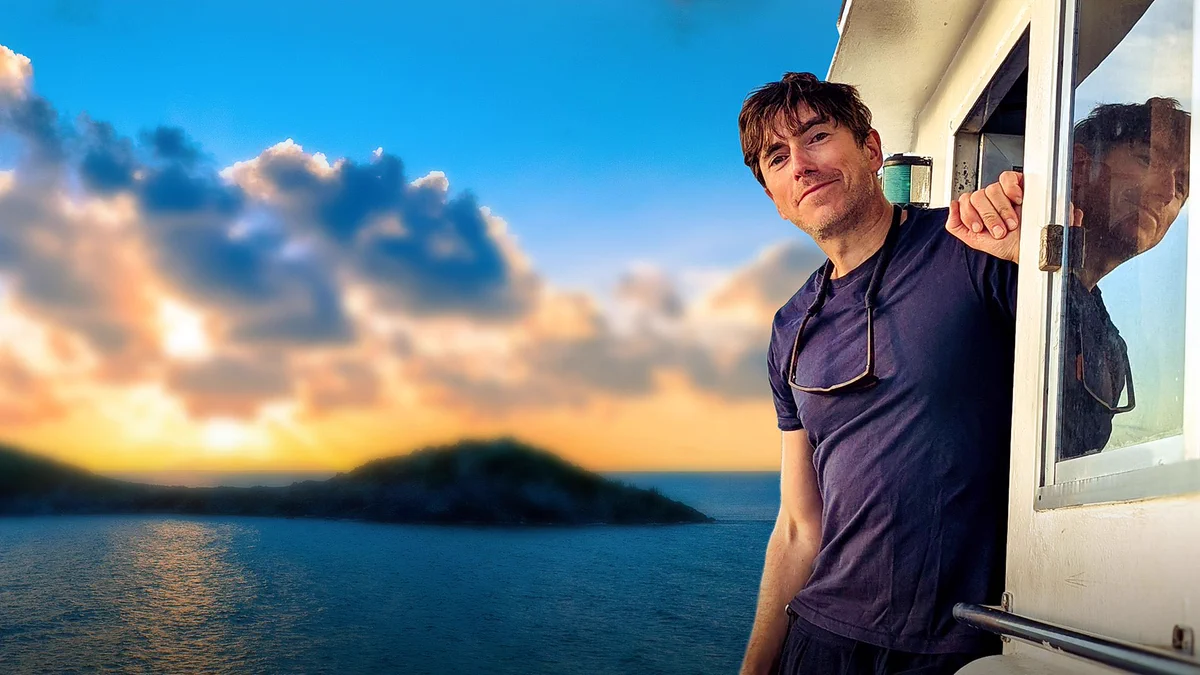 A man in a dark t-shirt smiles to camera as he leans out a door, set against a backdrop of islands in a vast blue sea. The sun is just visible through bright, fluffy clouds on the horizon.