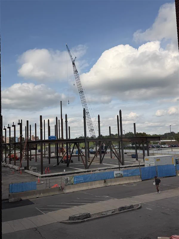 Construction site with a partially built steel building framework under a blue sky with white clouds. A tall crane is visible, and blue barriers delineate the site. 