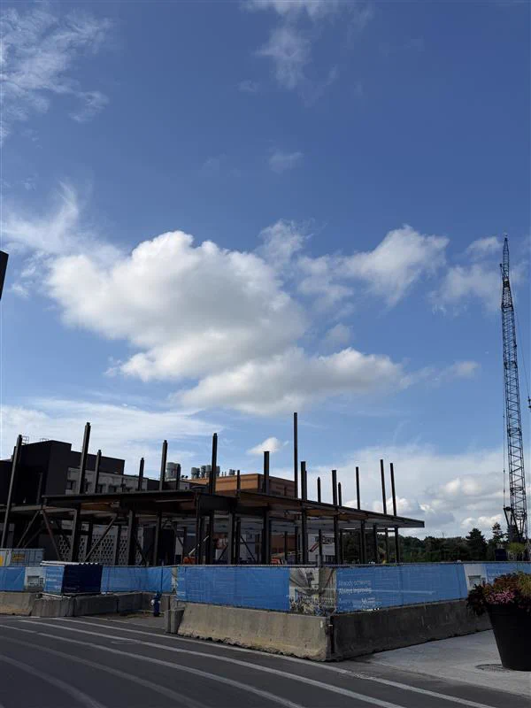 Steel framework of a building under construction, surrounded by concrete barriers and blue fencing displaying 'Always achieving. Always innovating.' A large construction crane rises on the right. Blue sky with white clouds above a paved road in the foreground.
